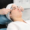 Woman laying on bed wearing sheet mask while esthetician uses silver ball massager on temples